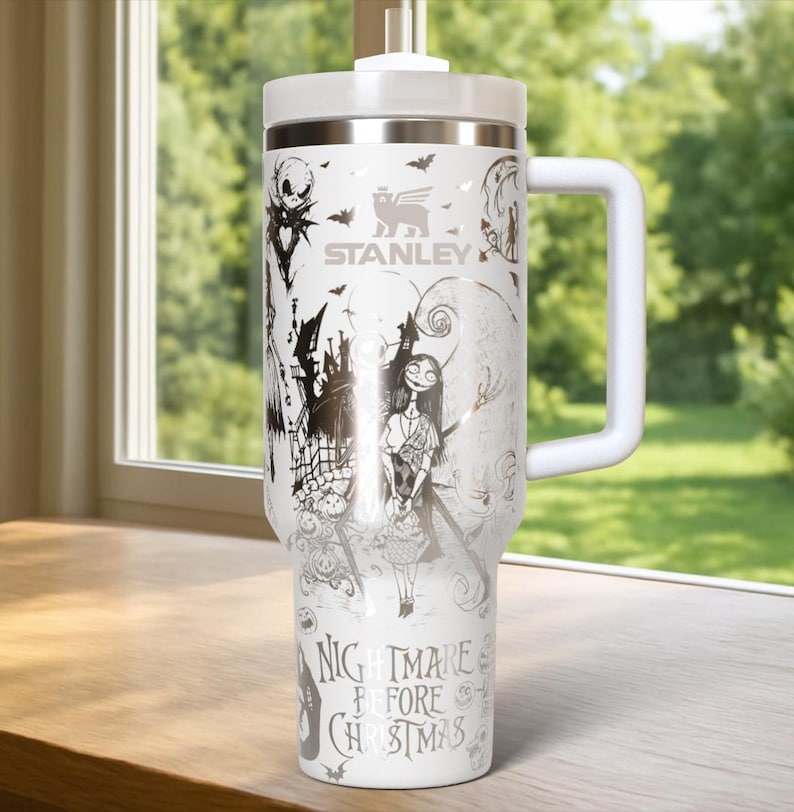 Puede incluir: Vaso Stanley blanco con un dise&ntilde;o de "Pesadilla antes de Navidad". El vaso tiene un asa y tapa blancos, y un gr&aacute;fico plateado de Jack Skellington, Sally y otros personajes. El texto "NIGHTMARE BEFORE CHRISTMAS" est&aacute; impreso en la parte inferior.