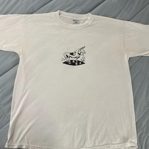 May include: White t-shirt with a black and white illustration of a unicorn-like creature with a spotted pattern, standing on a patch of grass with flowers. The shirt has short sleeves and a crew neck.