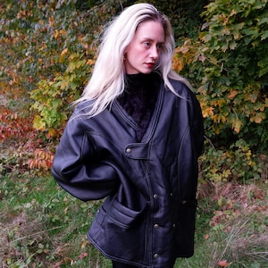 Pode incluir: Casaco de couro preto com gola de pele sintética e bolsos grandes. O casaco tem fecho de botão e uma silhueta clássica e oversized. A modelo usa calças pretas e está em frente a um fundo de folhagem verde e amarela.