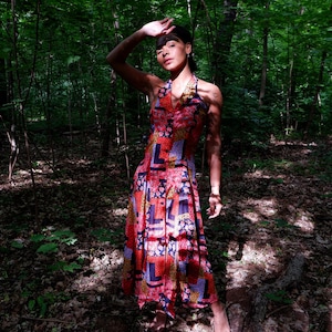 Può includere: Abito con collo all'americana con un vivace motivo floreale e geometrico in rosso, blu e giallo. L'abito ha un corpetto abbottonato e una gonna fluida. L'immagine è scattata all'aperto in un ambiente forestale.