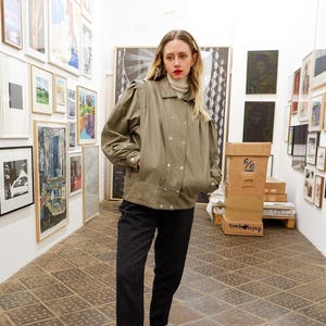 Pode incluir: Uma mulher vestindo uma jaqueta verde cáqui com gola alta e calças pretas. Ela está em uma galeria com obras de arte nas paredes.