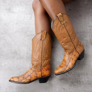 Könnte beinhalten: Ein Paar braune Cowboystiefel aus Leder mit einem strukturierten Schlangenledermuster im unteren Bereich und einem glatten Lederschaft. Die Stiefel haben eine spitz zulaufende Kappe, einen abgesetzten Blockabsatz und Ziernähte an den Schaftseiten.