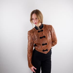 Könnte beinhalten: Eine kurze, hellbraune Lederjacke mit rundem Halsausschnitt und vier schwarzen, dekorativen Knebelverschlüssen auf der Vorderseite. Die Jacke besteht aus mehreren vernähten Lederbahnen und hat lange Ärmel mit einer taillierten Passform.