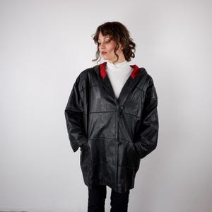 Op de afbeelding: Een zwart leren jack met een paneelconstructie, een capuchon met een contrasterende rode binnenvoering, lange mouwen en een knoopsluiting aan de voorzijde. Het jack wordt gedragen over een wit shirt met col en een zwarte broek.