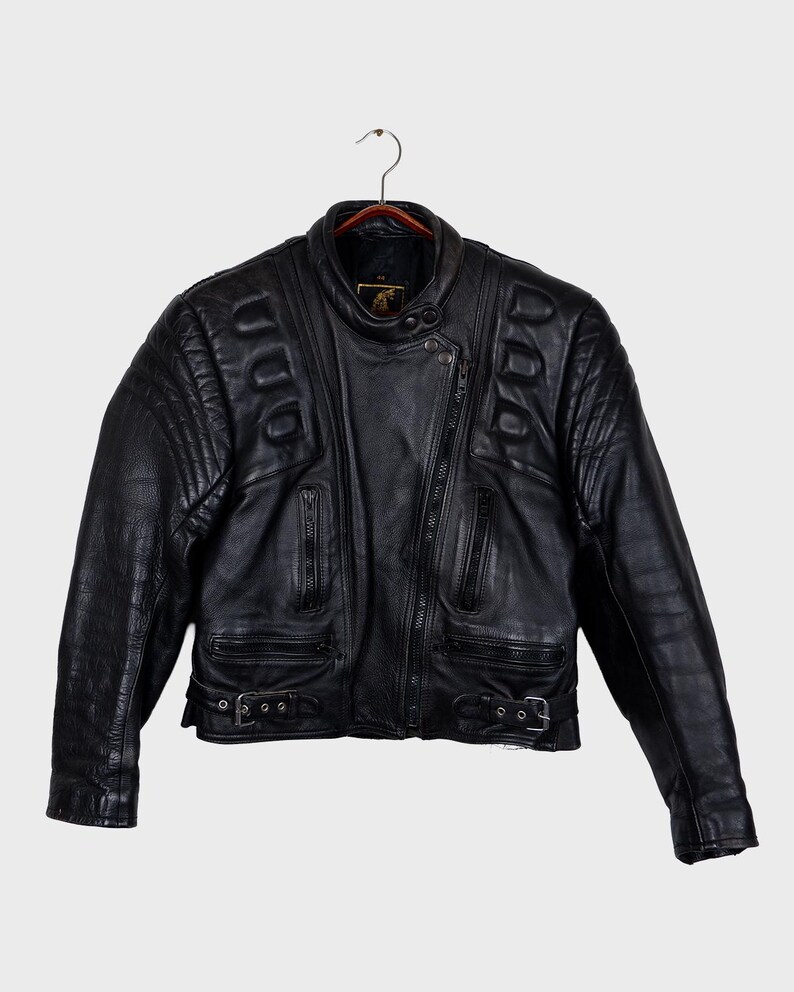 May include: Black leather motorcycle jacket with quilted shoulder and upper arm detailing. It has a diagonal zip, multiple zipped pockets, and adjustable waist straps with buckles. The collar is a stand-up style with a snap closure.