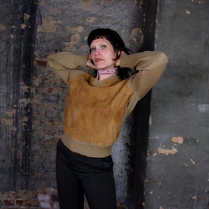 May include: A tan suede and knit sweater with a V-neck. The sweater has a suede front panel and ribbed knit sleeves. The person is wearing black pants and a patterned scarf. The background is a textured, gray wall.