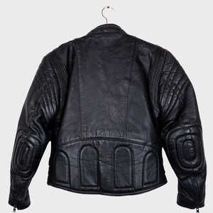 May include: Black leather motorcycle jacket with quilted shoulder and elbow padding. The jacket features a classic design with a stand-up collar, zippered cuffs, and arched details at the lower back. It hangs on a metal hanger.