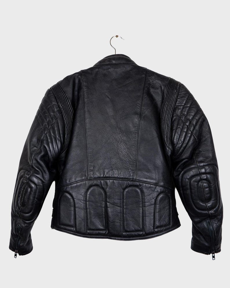 May include: Black leather motorcycle jacket with quilted shoulder and elbow padding. The jacket features a classic design with a stand-up collar, zippered cuffs, and arched details at the lower back. It hangs on a metal hanger.