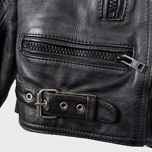May include: Close-up of a black leather jacket. The jacket has a zippered pocket, a buckle belt, and a zipper closure. The leather has a textured appearance.