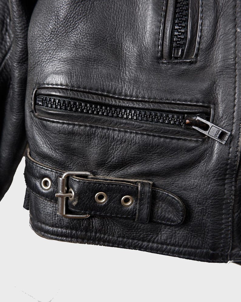 May include: Close-up of a black leather jacket. The jacket has a zippered pocket, a buckle belt, and a zipper closure. The leather has a textured appearance.