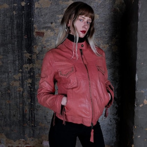 May include: A coral leather jacket with a zip-up front, pockets, and decorative studs. The jacket features a stand-up collar and tassel details. The model is wearing a black turtleneck and black trousers.