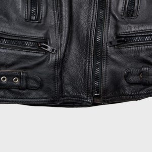 May include: Close-up of a black leather jacket with multiple zipped pockets and a central zip. The jacket has buckle straps and textured leather, typical of a classic biker style.