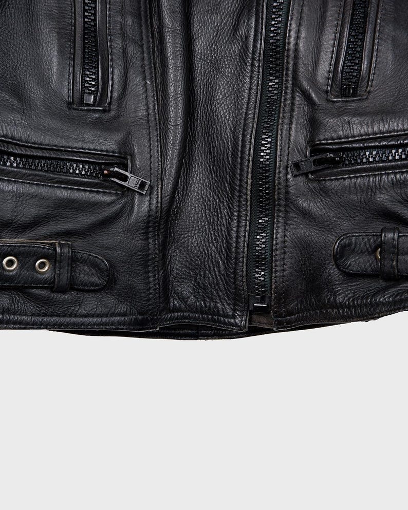 May include: Close-up of a black leather jacket with multiple zipped pockets and a central zip. The jacket has buckle straps and textured leather, typical of a classic biker style.