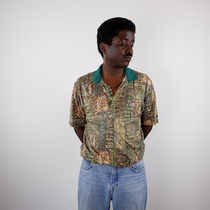 May include: A short-sleeved polo shirt featuring a multi-coloured abstract pattern in shades of green, brown, and tan, complemented by a solid dark green collar and a buttoned placket, paired with light blue denim jeans.