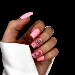 Op de afbeelding: Een hand met roze kunstnagels. De nagels zijn effen lichtroze, met twee accentnagels met vlinderontwerpen en glitter. De achtergrond is zwart.