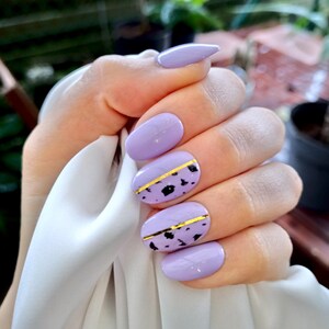 Op de afbeelding: Een hand met lavendelkleurige vingernagels. Twee nagels hebben een gouden streep met zwarte spikkels. De andere nagels zijn effen lavendel. De nagels zijn ovaal van vorm, met één puntige nagel. De hand houdt een witte doek vast.