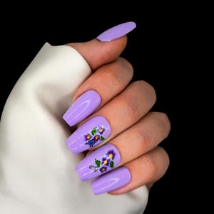 Op de afbeelding: Kunstnagels in lavendelkleur met een glanzende afwerking. Twee nagels zijn versierd met een bloemmotief in paars, geel en groen. De nagels zijn lang en rechthoekig, met een glad, egaal oppervlak.