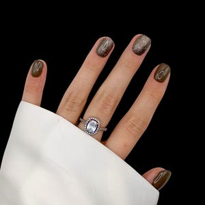 Op de afbeelding: Een hand met een zilveren ring met een vierkante, heldere steen omringd door kleinere stenen. De nagels zijn donker olijfgroen gelakt, sommige met glinsterende bruine nagellak. De achtergrond is effen zwart.
