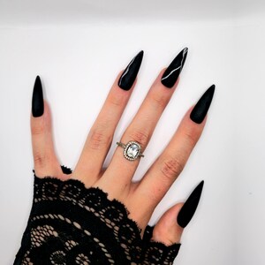 Op de afbeelding: Een hand met lange, zwarte, puntige vingernagels, één met een witte streep. Een zilveren ring met een vierkante, heldere steen en een halo van kleinere stenen zit aan de ringvinger. De hand is versierd met een zwarte kanten handschoen.