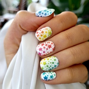 Op de afbeelding: Close-up van een hand met gelakte nagels. Elke nagel heeft een witte basis met een kleurovergang en een patroon van kleine sterren. De kleuren zijn onder meer blauw, groen, geel, oranje en rood.