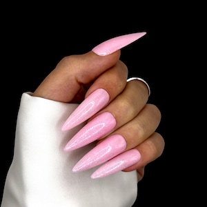 Op de afbeelding: Close-up van lange, puntige, lichtroze glitter nagels. De nagels hebben een effen kleur met een sprankelende afwerking. De hand is tegen een zwarte achtergrond.