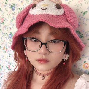 May include: A pink crocheted hat with a white character design on the front. The hat is worn by a person with long, red hair and black framed glasses. The person is wearing a pink choker necklace with a heart pendant.