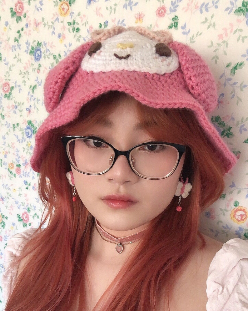May include: A pink crocheted hat with a white character design on the front. The hat is worn by a person with long, red hair and black framed glasses. The person is wearing a pink choker necklace with a heart pendant.