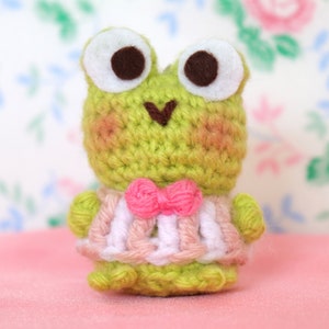 May include: A green crocheted frog wearing a pink and white striped dress with a pink bow. The frog has large brown eyes and a heart-shaped mouth.