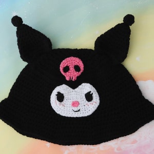 May include: Black crocheted hat with a white and pink skull and crossbones design. The hat features a white crocheted face with black eyes and a pink nose.