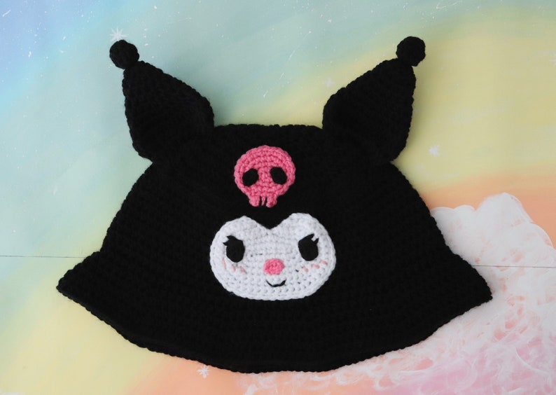 May include: Black crocheted hat with a white and pink skull and crossbones design. The hat features a white crocheted face with black eyes and a pink nose.