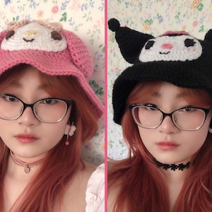 May include: Two women with red hair are wearing crocheted hats. The hat on the left is pink with a white character on the front. The hat on the right is black with a white character on the front. Both women are wearing glasses.