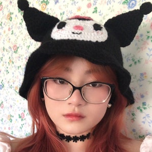 May include: A person with long, pink hair is wearing a black crocheted hat with white and pink cartoon character details. The hat has large, floppy ears. The person is also wearing black framed glasses and a black choker necklace.