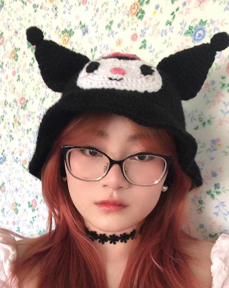 May include: A person with long, pink hair is wearing a black crocheted hat with white and pink cartoon character details. The hat has large, floppy ears. The person is also wearing black framed glasses and a black choker necklace.