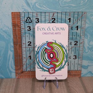 Puede incluir: Un pin colorido con un diseño único, con círculos concéntricos rojos, verdes, azules y blancos y una forma roja estilizada. El pin está en una tarjeta blanca con el texto "Fox & Crow CREATIVE ARTS".