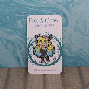 Puede incluir: Un pin con un personaje de dibujos animados con pelo amarillo, un sombrero rosa y un atuendo negro. El pin está sobre una tarjeta blanca con el texto "Fox & Crow CREATIVE ARTS" y el identificador "@foxandcrowcreative". El fondo es un patrón veteado azul y turquesa.