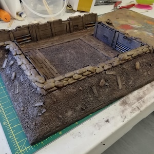 Puede incluir: Un modelo en miniatura de una trinchera con un marco de madera y paredes de piedra. La trinchera está hecha de materiales marrones y grises y está colocada sobre una estera de corte verde.