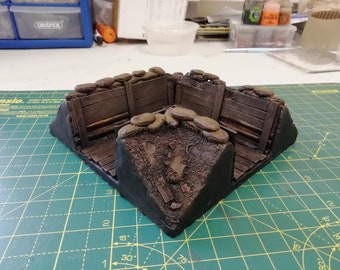 May include: A miniature model of a trench with wooden walls and sandbags. The trench is made of brown and black materials and is set on a green cutting mat.
