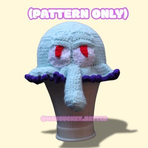 Puede incluir: Un gorro de ganchillo azul claro que se asemeja a Calamardo de Bob Esponja, con ojos blancos, pupilas rojas y una nariz larga. El gorro tiene un ribete morado y el texto "(PATTERN ONLY)".