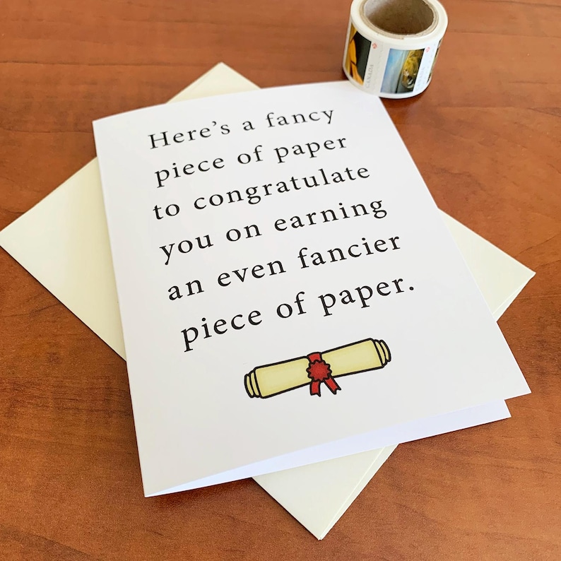May include: A white greeting card with the text "Here's a fancy piece of paper to congratulate you on earning an even fancier piece of paper." A scroll illustration is at the bottom. The card is on a cream envelope.