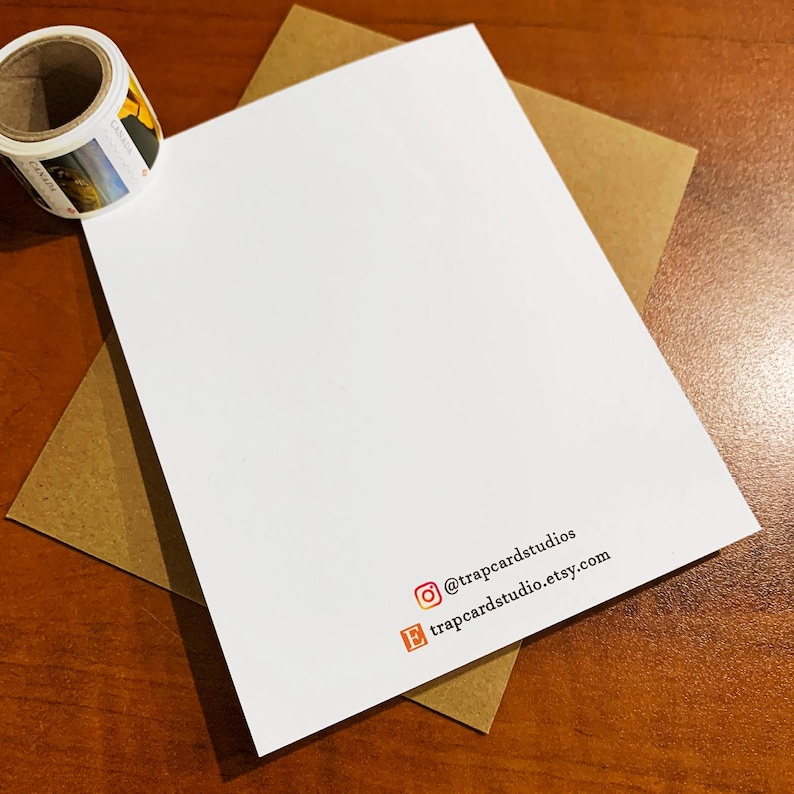 Pode incluir: Um cart&atilde;o branco com o texto "@trapcardstudios" e "trapcardstudio.etsy.com" impresso nele. O cart&atilde;o est&aacute; sobre uma superf&iacute;cie marrom.