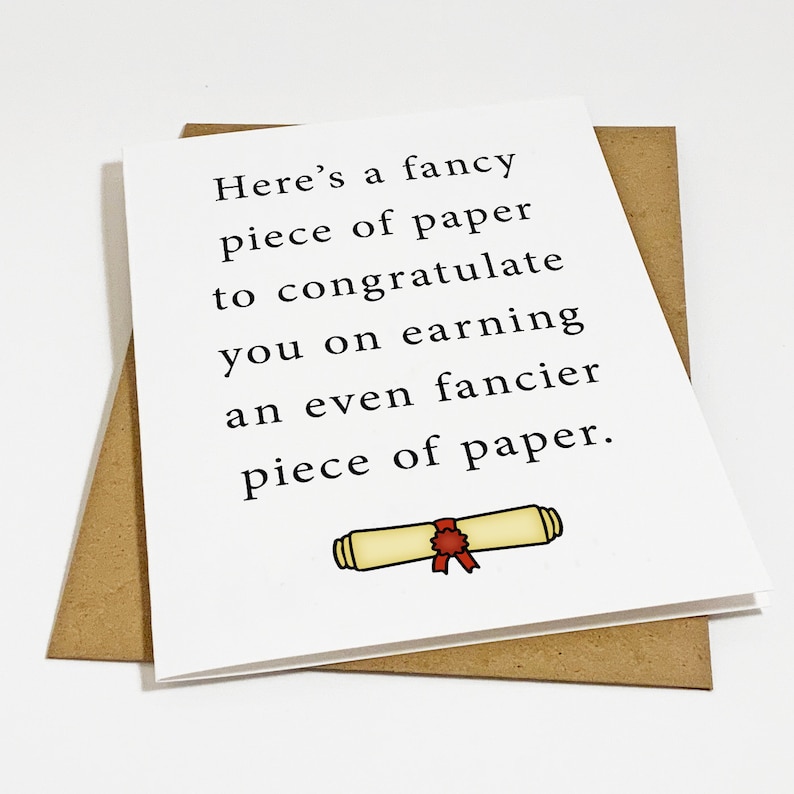 May include: A white graduation card with black text that reads, "Here's a fancy piece of paper to congratulate you on earning an even fancier piece of paper." A cartoon scroll illustration is at the bottom. The card is on a brown envelope.