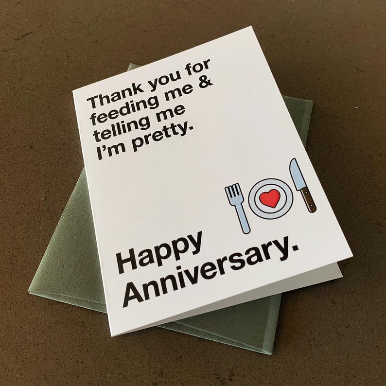 Puede incluir: Una tarjeta blanca con texto negro que dice "Gracias por alimentarme y decirme que soy bonita. Feliz aniversario." Una ilustraci&oacute;n simple de un plato, un tenedor y un cuchillo con un coraz&oacute;n rojo en el plato est&aacute; debajo del texto.