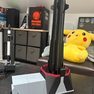 May include: A black and gray model rocket with a red platform and a gray launch structure. The rocket has a pointed nose cone and fins. A smaller black and white rocket model is also visible.
