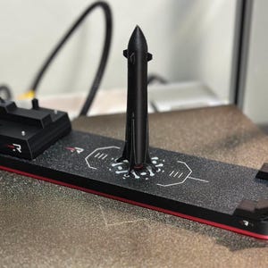 Pode incluir: Modelo preto impresso em 3D de um foguete e plataforma de lançamento. O foguete é alto e esguio, com um nariz pontudo. A plataforma é retangular, com superfície preta e borda vermelha. O modelo está sobre uma superfície texturizada.