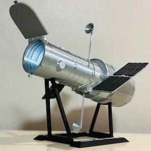 May include: A silver model of the Hubble Space Telescope, featuring a cylindrical body with a blue interior, solar panels, and a support stand. The model is detailed with various components and a gray protective cover.