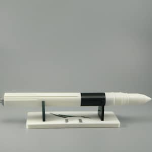 May include: A white and black model rocket on a white display stand. The rocket has a black band around the middle and a white pointed nose cone. The stand has the letter "F" printed on it.