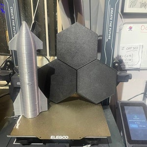 May include: A 3D printed model of a rocket ship on a 3D printer bed. The printer is black and has a white label that says "ELEGOO". The printer is also labeled "CREATE THE FUTURE".
