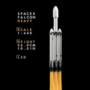 May include: A white SpaceX Falcon Heavy rocket model with black detailing and orange LED light exhaust flames. Text reads: SPACEX FALCON HEAVY, SCALE 1:440, HEIGHT 26.0 CM, 10.0 IN, LED. The rocket features three cylindrical boosters and a central payload fairing.