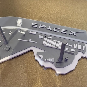 May include: A gray and white SpaceX logo with the word SPACEX in capital letters. The logo has two gray, cylindrical structures with a screw design. The logo is a 3D printed item.