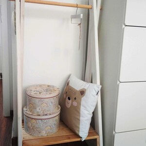 May include: White wooden clothing rack with a wooden bar, two round storage boxes with floral patterns, and a white pillow with a brown bear face design.